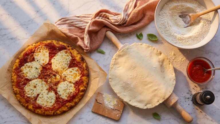 Golden gluten-free pizza crust with cheese and tomato sauce beside GF flour and dough
